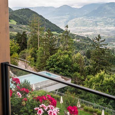 Blick von einem Balkon mit blühenden roten Geranien auf einen modernen Swimmingpool inmitten einer bewaldeten Hügellandschaft mit einem Tal im Hintergrund.