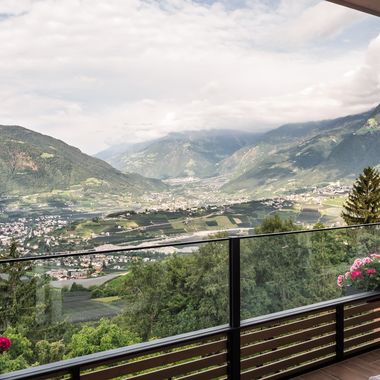 Ein Blick von einem Balkon mit Glasgeländer und bepflanzten Blumenkästen auf ein weites Tal mit einer Stadt und umliegenden Bergen bei bewölktem Himmel.