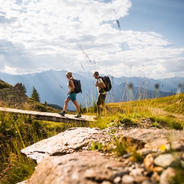 Ein Paar mit Wanderrucksäcken geht über eine Holzbrücke in einer bergigen Landschaft bei sonnigem Wetter.