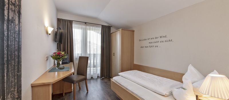 Camera singola con letto, armadio in legno chiaro, scrivania con sedia e finestra con tende.