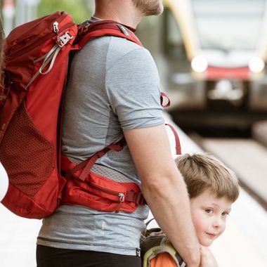 Eine Familie mit Rucksäcken steht auf einem Bahnsteig und wartet auf einen einfahrenden Zug.