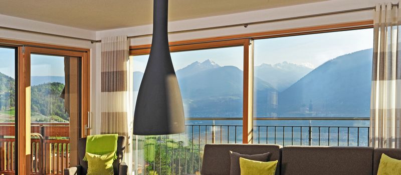 Soggiorno con grandi finestre che mostrano vista su montagne e valle, mobili moderni con cuscini verdi.