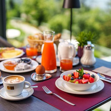 Ein reich gedeckter Frühstückstisch auf einer Terrasse mit einer Schüssel frischem Obstsalat, zwei gekochten Eiern, Tassen mit Cappuccino, Orangensaft und verschiedenen Brotaufstrichen.