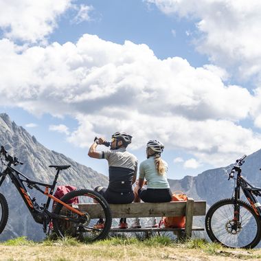 Ein Ehepaar in Fahrradkleidung und mit Helmen sitzt auf einer Holzbank mit Blick auf ein Bergpanorama, ihre E-Bikes sind links und rechts neben der Bank geparkt.