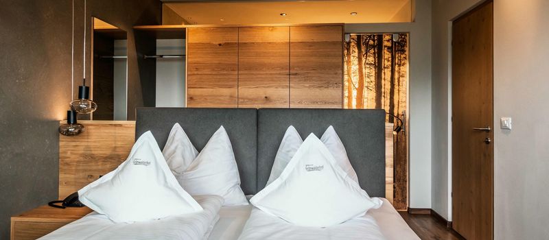 Camera d'albergo moderna con due letti singoli affiancati, testiera grigia, cuscini bianchi e armadio in legno sullo sfondo.