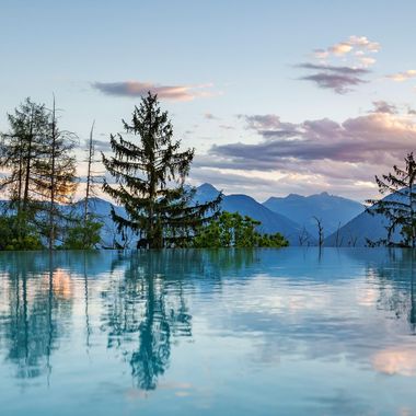 Ein Infinity-Pool spiegelt bei Abenddämmerung die umliegenden Tannen und das ferne Gebirgspanorama wider.