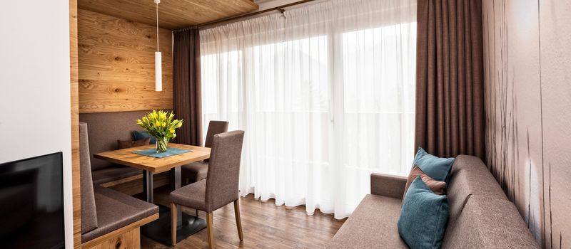 Soggiorno moderno con divano marrone, tavolo in legno con fiori gialli e ampie tende bianche e scure.