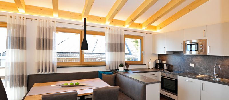 Cucina moderna con soffitto a travi di legno, tavolo da pranzo con sedie e divanetto, grande finestra e elettrodomestici integrati.