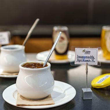 Ein weißes Porzellanschälchen mit Aprikosenmarmelade und einem Silberlöffel steht auf einem dunklen Tisch, daneben ein Kleinschild mit der Aufschrift 'hausgemachte Aprikosen M.'.